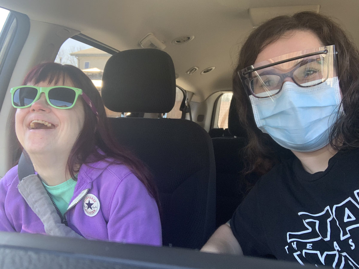 A DSP wearing personal protective equipment and  the person they support laugh together in a car. 